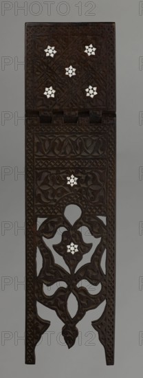 Quran lectern rahla, Islamic art, Turkish, Ottoman dynasty 1299–1922; Quran lectern decorated with arabesques, geometric ornament, lotuses, tulips, rosettes; 19th century, Turkey; piece of furniture; unfolded: height 50.6 cm, width 91.5 cm, depth 21.3 cm, weight 1.7 kg; folded: height 80.5 cm, width 21.3 cm, weight 1.7 kg., A vertically oriented carved wooden panel with dark brown color featuring intricate swirling and interlaced vegetal motifs, openwork sections, and several small white floral inlays arranged in star-like patterns; the top portion is a rectangular hinged element with carved borders and a few white inlaid stars, below which a carved band with geometric and scrollwork patterns leads to an open lattice area with ornate curving shapes and another white inlaid star, all set against a neutral gray background.