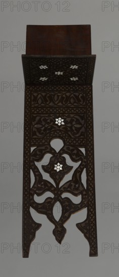 Quran lectern rahla, Islamic art, Turkish, Ottoman dynasty 1299–1922; Quran; decorated with arabesques, geometric ornament, lotuses, tulips, rosettes; 19th century, Turkey; piece of furniture; unfolded: height 50.6 cm, width 91.5 cm, depth 21.3 cm, weight 1.7 kg; folded: height 80.5 cm, width 21.3 cm, weight 1.7 kg., The image shows a tall, narrow carved wooden panel with ornate openwork and inlaid geometric flower shapes. The wood is dark brown with lighter brown highlights along carved edges, and there are several small white inlaid flower motifs set into the carved patterns. The panel features a rectangular top piece followed by a section of dense carved vines and scrolls, a central area with an open, symmetrical floral motif, and a lower tapering section with interlaced curves and pointed tips. The background behind the panel is a flat gray color.