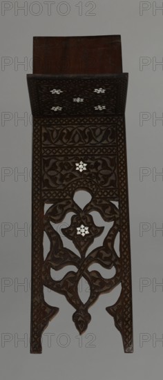 Quran lectern rahla, Islamic art, Turkish Ottoman dynasty, 19th century. Wood lectern for Quran, decorated with arabesques, geometric ornaments, rosettes, lotus/tulip motifs. Function: piece of furniture. Dimensions unfolded: H 50.6 cm, W 91.5 cm, D 21.3 cm, weight 1.7 kg; folded: H 80.5 cm, W 21.3 cm, weight 1.7 kg., A tall carved wooden object in dark brown fills the image against a light gray background, showing an upper flat panel and a long vertical section with ornate openwork carvings; the carvings form flowing vine- and leaf-like shapes and geometric borders, and there are a few small inlaid light-colored floral motifs set within the carved patterns, creating contrast with the dark brown wood.