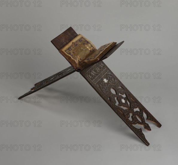 Quran lectern rahla, Islamic art, Turkish, Ottoman dynasty 1299–1922; Quran lectern decorated with arabesques, geometric ornament, rosettes, lotus tulip motifs; 19th century, Turkey; piece of furniture; unfolded: height 50.6 cm, width 91.5 cm, depth 21.3 cm, weight 1.7 kg; folded: height 80.5 cm, width 21.3 cm, weight 1.7 kg., A carved wooden bookrest with ornate lattice-like cutouts and floral motifs holds an open illuminated book; the bookrest and book covers are dark brown wood, the carved panels show lighter brown highlights, and the carved inlay appears pale; the open book has warm yellowed pages with gold and colored ornamentation in tones of gold, deep blue, red, and green; the background is a smooth neutral gray, and soft shadows appear beneath the bookrest.
