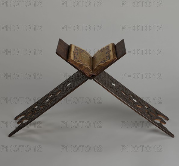 Quran lectern rahla, Ottoman Turkey, 19th century. Wooden piece of furniture for holding the Quran, decorated with arabesques, geometric ornaments, rosettes, lotus tulip motifs, lotuses and tulips. Inscription/classification details not specified. Unfolded: height 50.6 cm, width 91.5 cm, depth 21.3 cm, weight 1.7 kg. Folded: height 80.5 cm, width 21.3 cm, weight 1.7 kg., A carved wooden bookrest holding an open book, positioned with its two dark brown wooden panels crossed in an X shape; the bookrest has intricate cut-out and carved patterns and lighter inlaid accents, with tones of dark brown and hints of lighter wood; the open book shows aged pages with decorative colored borders and patterns in shades of gold, brown, blue, and red, and the book’s cover appears darker brown; the background is a smooth neutral gray.