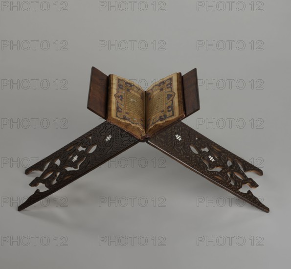 Quran lectern rahla, Islamic art, Turkish, Ottoman dynasty 1299–1922. Quran lectern decorated with arabesques, geometric ornament, rosettes, lotus tulip motif; decorated with rosettes. 19th century, Turkey. Piece of furniture. Dimensions unfolded: height 50.6 cm, width 91.5 cm, depth 21.3 cm, weight 1.7 kg; folded: height 80.5 cm, width 21.3 cm, weight 1.7 kg., An open book with ornate patterned page edges rests on a carved wooden book stand; the book's pages show intricate decoration in shades of gold, blue, red, and brown, and the covers are dark brown; the stand is dark wood with carved, interlacing floral and geometric cutouts and small inlaid lighter-colored accents, set against a neutral light gray background.