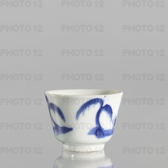 Bowl, ceramics, blue-and-white porcelain, Qing 1644–1911, 19th century, China. Bowl with flared edges on a low ring foot; grayish-white porcelain decorated with landscapes with architecture and staffage. Height 4.4 cm, diameter 5.8 cm.,  noted., A small ceramic cup sits on a reflective surface against a light gray background, the cup is white with a glossy finish and decorated with a few blue brushstroke motifs that resemble curved shapes and leaflike forms, the rim of the cup is slightly flared and the base is a short foot with a subtle ring where it meets the surface, visible colors include white, blue, light gray, and a hint of off-white on the base.