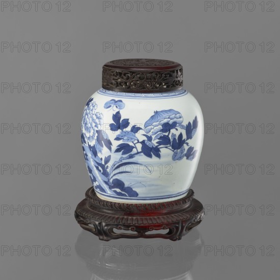 Storage pot on a stand with a cover, Chinese, Qing dynasty 1644–1911, 19th century. Blue-and-white porcelain storage pot decorated with butterflies, peonies, and floral motifs. Cover b round, decorated throughout with openwork peony flowers and tendrils. Stand c round, shallow, recessed with a central round recess; decorated with a tongue ornament and openwork. Vessel; height 19.0 cm, diameter 19.5 cm, weight 2.3 kg., A rounded ceramic jar with a carved dark wooden lid sits on an ornate dark wooden stand; the jar has a white background with blue painted floral and foliage designs, including large blossoms and leaves, and a decorative blue band near the top, while the wooden lid and stand show rich brown and reddish-brown tones.