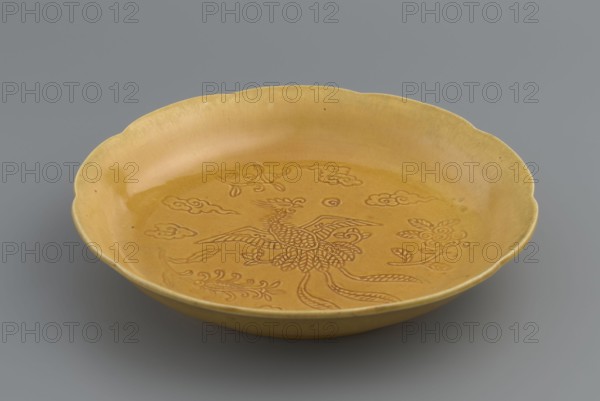Plate, Paderewski, Ignacy Jan 1860-1941 provenance: gift, Chinese, Qing dynasty 1644–1911, 18th–19th century. Monochromatic yellow-glazed vessel, engraved decoration: phoenix among clouds and plant ornament. Height 3 cm; diameter 16.5 cm., A shallow round dish with a scalloped rim, glazed in a warm yellow color, sits against a neutral gray background; the interior shows an incised decorative design featuring a stylized bird surrounded by floral and cloud-like motifs in the same yellow tone with slightly darker engraved lines, and the dish surface displays subtle variations of yellow and small darker specks.