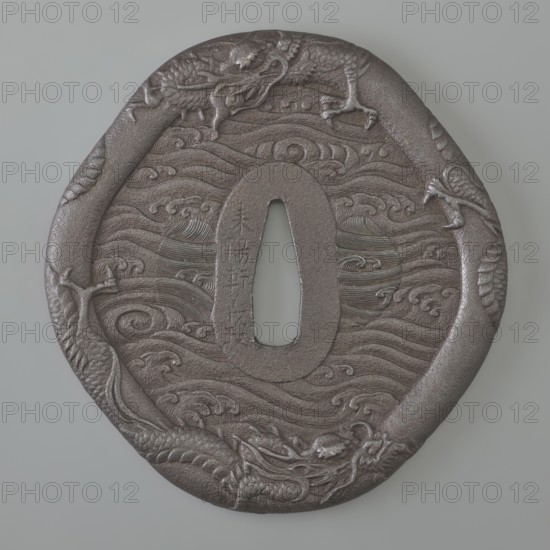 Crossguard tsuba, Raiyôken, Yoshimasa maker, sword crossguard in the shape of an otafuku chubby woman's face. Edo period, Kaei 5 1852, Year of the Rat, Japan. Iron tsuba, height 8.7 cm, width 8.35 cm, depth 0.5 cm. Edge thick, clearly defined and rounded hira dote mimi; oval seppa dai. Decorated in low relief with two dragons; shield guard surfaces carved on both sides in low relief with rough sea waves umiji. Outlines of the side hitsu holes visible, later plugged with different engraving style. Edge decoration includes two dragons. Inscription phrase kitsu jitsu happy day present. Weapons, katana; motifs: dragons, sea waves, rat zodiac; melee weapons; Japanese art; nô character Otafuku., A round, dark brown metal object with an oval hole near its center sits against a pale gray background; the surface shows carved swirling wave patterns and a raised border that forms a continuous serpentine dragon motif with textured scales and a detailed dragon head visible along the lower edge, and...