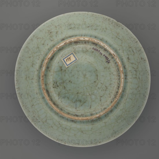 Plate, Chinese, Qing dynasty 1644–1911, 18th century, porcelain vessel with inscriptions, lotus and plant decoration; height 3.5 cm, diameter 21.6 cm, weight 0.5 kg., A circular plate viewed from above with a pale green glazed surface showing a network of visible crackle lines and scattered darker speckles, a recessed central area defined by a raised ring with exposed beige rim edges, a small decorative square label with a blue border adhered near the central area, and an overall color palette of pale green, beige, blue, and scattered brownish-gray specks against a neutral gray background.