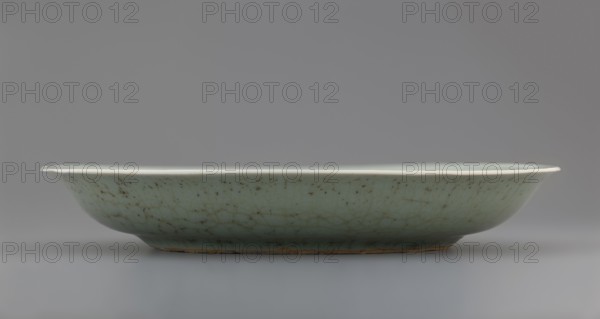 Plate, Chinese, Qing dynasty 1644–1911, 18th century, porcelain vessel with inscriptions and decoration of lotuses and plants; height 3.5 cm, diameter 21.6 cm, weight 0.5 kg., A shallow oval dish sits on a flat surface, viewed from the side, showing a pale green body with a network of fine darker crackle lines and some brown speckling and spots; the rim appears lighter, near white, and the base edge shows a thin band of exposed brown clay; the background and surface are shades of gray.