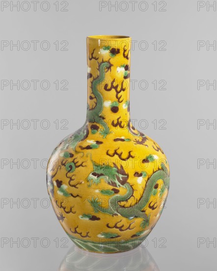 Vase, ; Chinese; Qing dynasty 1644–1911, Kangxi period 1662–1722; cylindrical-neck vessel with fa-hua decoration featuring dragons iconography; material: ceramic decorative vessel; date: 1662–1722; height 41.0 cm, diameter 24.3 cm, weight 3.0 kg., A tall, rounded ceramic vase with a long neck sits against a plain light gray background; the vase is predominantly bright yellow with a glossy finish and is decorated with a group of green dragons outlined in darker green and brown, surrounded by several cloud-like motifs in shades of green, white, and brown, plus numerous flame-like shapes in dark brown; the base of the vase has a band of layered wave patterns in green and white.
