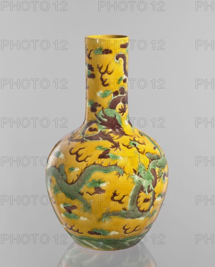 Vase, Chinese; Qing dynasty, Kangxi period 1662–1722; porcelain cylindrical-neck vessel with fa-hua decoration depicting dragons; height 41.0 cm, diameter 24.3 cm, weight 3.0 kg., A tall ceramic vase with a narrow neck and rounded body sits against a plain light gray background; the vase is predominantly yellow with decorative motifs in green, brown, and teal, featuring a group of sinuous dragon figures with scaled bodies and clawed limbs entwined around the surface, surrounded by stylized cloud-like shapes and flame-like elements, and a band of green wave patterns encircling the base; highlights and reflections on the glaze indicate a glossy finish.