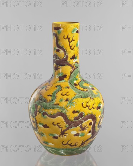 Vase, Chinese, Qing dynasty Kangxi, 1662–1722, fa-hua decoration, dragons iconography, cylindrical neck decorative vessel, porcelain, China, 1662–1722, height 41.0 cm, diameter 24.3 cm, weight 3.0 kg, A ceramic vase with a tall narrow neck and rounded body decorated in bright yellow glaze, featuring several sinuous dragons in green and dark brown with textured scale patterns, surrounded by cloudlike motifs in green, white, and dark brown, and areas of darker brown flame shapes; the base has a band of green wave patterns and the surface shows glossy reflections against a neutral gray background.
