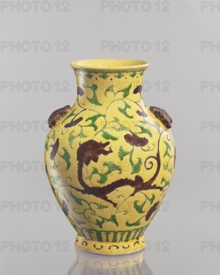 Vase. Chinese; Qing dynasty 1644–1911, 19th century. Porcelain vessel, height 24.5 cm, diameter 17.5 cm, weight 1.35 kg. Yellow glaze with engraved decoration, partly applied plastic elements, painted with green and brown enamels. Low foot; neck cup-shaped toward the rim. Two applied plastic butterflies on the belly arms. Motifs: butterflies and plant ornaments., A ceramic vase with a yellow ground and a bulbous body tapering to a narrower neck and foot, decorated with flowing green vine and leaf motifs and dark brown floral or foliate shapes; there are a few applied dark brown decorative elements on the shoulder, a molded green band around the foot, and decorative green and brown details around the rim; the surface shows a glossy finish and some subtle darker speckling against the yellow background, and the vase sits on a reflective gray surface with a neutral pale gray background.