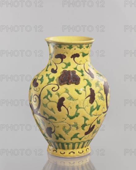 Vase., Chinese, Qing dynasty 1644–1911. Ceramic vessel, 19th century. With a low foot; neck cup-shaped toward the rim. Two applied plastic butterflies on the shoulders of the belly. Yellow glaze with engraved decoration, partly modeled plastic, painted with green and brown enamel. Height 24.5 cm; diameter 17.5 cm; weight 1.35 kg., A yellow ceramic vase with a flared rim and bulbous body decorated with a flowing pattern of green leafy tendrils and dark brown stylized floral or cloud-like motifs, with a green and yellow band around the base and additional green decorative accents near the neck, set against a neutral light gray background.