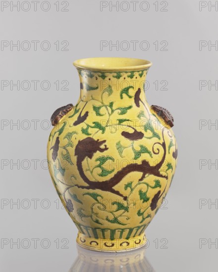 Vase. Chinese; Qing dynasty 1644–1911, 19th century. Porcelain vessel, height 24.5 cm, diameter 17.5 cm, weight 1.35 kg. Yellow glaze with engraved decoration, partly modeled plastic elements, painted with green and brown enamel. With a low foot and a cup-shaped neck toward the rim. Two applied plastic butterflies on the sides of the belly. Motifs: butterflies and plant ornaments., A yellow ceramic vase with a flared rim and bulbous body sits against a pale gray background, decorated with a pattern of green scrolling vines and leaves and dark brown abstract floral or foliate shapes; there are a few raised brown ornamental handles on the neck area, a green decorative band near the base, and subtle reflections beneath the vase.