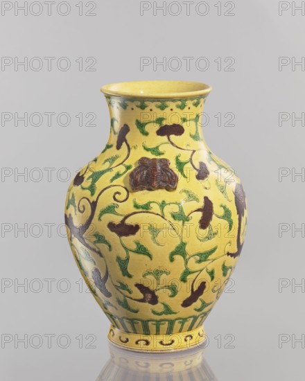 Vase., Chinese; Qing dynasty 1644–1911. Ceramic vessel, 19th century. With a low foot and a cup-shaped neck toward the rim. On the sides of the belly are two modeled plastic butterflies. Yellow glaze with engraved decoration, partly modeled, painted with green and brown enamel paints. Height 24.5 cm; diameter 17.5 cm; weight 1.35 kg. Subjects: butterflies and plant ornaments., A yellow ceramic vase with a rounded body, a narrow neck that flares at the rim, and a short foot, decorated with a repeating pattern of green scrolling vines and leaves and dark brown stylized floral or cloud shapes; there is a raised dark brown ornamental motif on the front, a green band of vertical elements above the foot, and a decorative border near the rim; the surface shows a glossy finish and subtle speckling, and the vase sits on a reflective pale surface against a light gray background.