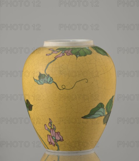 Ginger vessel with lid, Nagoya Shippô Kaisha c. 1870–1880, Meiji period 1868–1912. Small ovoid porcelain vase with round flattened lid; exterior covered with lemon-yellow glaze with decorative crackle grid, interior and base white with transparent glaze. Decorated with a naturalistic bean-vine motif of greenish-brown leaves and pink flowers, loosely twining around the body and reaching the lid. Motif executed in cloisonné enamel using brass wires and enamel in several shades of green and brown; the design was carved into the vessel surface before filling and firing totai-shippo technique. Signature in underglaze cobalt: Nihon Shippo Kaisha sei zo; overglaze red-iron stamp in a rhomboid cartouche with the sign so presumed exporter’s mark. Porcelain cloisonné developed around 1868 by Tsukamoto Kaisuke 1828–1887; similar pieces were made by Takeuchi Chubei and Suzuki Seiichiro, who worked for Nihon Shippo Kaisha in Nagoya. Height 12.4 cm; diameter 9.6 cm., A rounded ceramic vase with a ye...