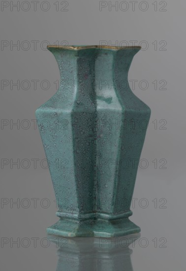 Vase, ; Qing style; ceramics; Chinese art; Chinese culture; Qing dynasty 1644–1911; 19th century, China; vessel; height 11.6 cm, width 4.7 cm, depth 7.15 cm., A ceramic vase with an angular, faceted silhouette sits against a neutral background, displaying a predominantly turquoise-green glaze with a mottled, speckled surface pattern and subtle darker green and bluish variations; the rim shows a thin band of muted gold, the vase tapers inward toward the neck then flares slightly at the top and narrows again above a stepped base, and the base sits on a reflective surface that creates a soft mirrored reflection beneath it.