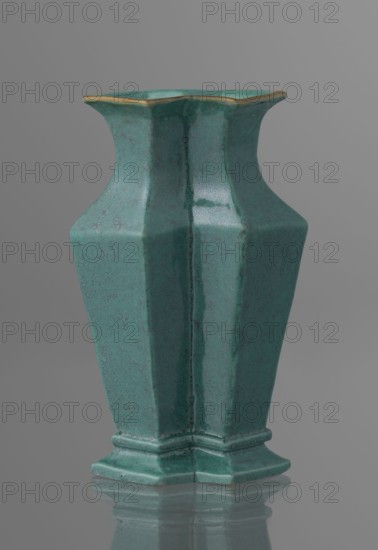 Vase;, ; Chinese; Qing dynasty 1644–1911; 19th century; ceramics; covered with a turquoise glaze with manganese spots; vessel; height 11.6 cm, width 4.9 cm, depth 7.1 cm, weight 0.15 kg., A ceramic vase with a tall, angular shape and a flared rim sits against a smooth gray background, the vase surface showing a muted turquoise-green color with subtle darker speckling and a slightly glossy finish, a thin metallic gold trim along the edge of the rim, a faceted body that narrows at the neck and widens toward a stepped base, and a soft reflection of the vase visible on the surface beneath it.