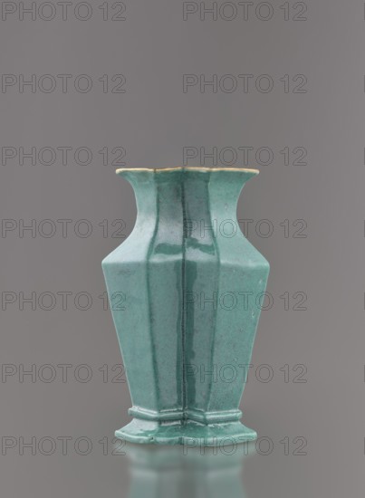 Vase, Chinese, Qing dynasty 1644–1911, 19th century. Ceramic vessel covered with a turquoise glaze with manganese spots. Height 11.6 cm; width 4.9 cm; depth 7.1 cm; weight 0.15 kg., A ceramic vase with a bluish-green glaze stands against a plain gray background, showing a hexagonal, slightly flared rim with a thin band of a pale yellowish edge, a tapered faceted body with visible vertical seams and subtle darker streaks in the glaze, and a stepped hexagonal base with rounded edges; the glaze surface displays a mottled texture and some darker green pooling in areas, and the vase casts a soft reflection on the surface below.
