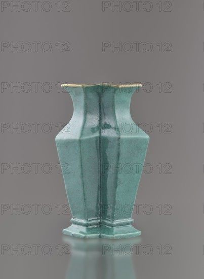 Vase, gift, Chinese art, Chinese culture, Qing 1644–1911, Qing style, ceramics, covered with a turquoise glaze with manganese spots, 19th century, China, vessel; height 11.6 cm, width 4.9 cm, depth 7.1 cm, weight 0.15 kg, A ceramic vase with a matte turquoise glaze standing on a reflective surface, featuring an angular, faceted body that tapers inward toward the neck and flares outward at the rim, where a thin band of muted beige outlines the edge; the surface shows subtle darker turquoise streaks and speckled texture, and the base has a stepped, hexagonal form with similar turquoise tones and faint glossy highlights, all set against a smooth gray background.