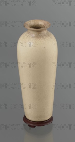 Cylindrical porcelain vase, tapering towards the bottom; flat rim. Light cream glaze with fine cracklework. Wooden, round, flat saucer on three legs. 19th century, Japan; Japanese, Edo 1603–1868. Ceramics; cracklework decoration. Vessel; height 21.1 cm, diameter 8.4 cm, weight 0.6 kg., A tall narrow vase with a rounded shoulder and flared rim sits on a small round base. The vase surface is a pale beige with a fine network of darker crackle lines and a few small dark spots, and the rim and interior show a slightly darker brownish-beige tone with more pronounced crackling. The base is a dark reddish-brown wood color and the background is a uniform medium gray.