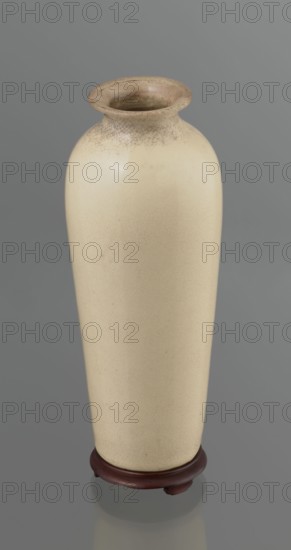 Cylindrical porcelain vase, tapering towards the bottom; flat rim. Light cream glaze with fine cracklework. Wooden, round, flat saucer on three legs. 19th century, Japan. Height 21.1 cm; diameter 8.4 cm; weight 0.6 kg., A tall, narrow ceramic vase with a rounded shoulder and a flared rim sits on a small wooden stand; the vase's surface is a pale cream color with subtle crackle texture near the rim and faint imperfections on the body, while the rim itself shows warmer beige and light brown tones; the wooden stand is a deep reddish-brown, and the background is a smooth medium gray.