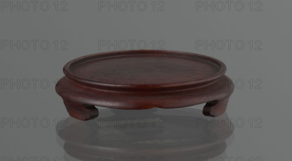 Vase on saucer. Cylindrical porcelain vase tapering toward the bottom with a flat rim. Light cream glaze with fine cracklework decoration. Wooden round flat saucer on three legs. 19th century, Japan Edo period. Ceramic vessel. Height 21.1 cm; diameter 8.4 cm; weight 0.6 kg., A low, round wooden stand with a shallow raised lip and curved sides rests on a reflective surface; the wood shows a deep reddish-brown color with darker brown grain variations and subtle worn spots, and the background and reflection appear gray.