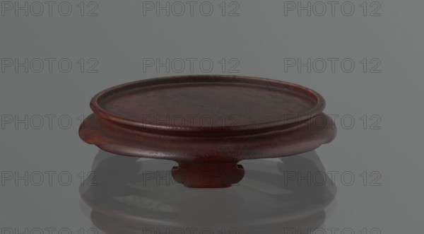 Vase on saucer. Cylindrical porcelain vase tapering toward the bottom with a flat rim; light cream glaze with fine cracklework. Wooden round flat saucer on three legs. 19th century, Japan; Edo 1603–1868. Japanese ceramics; saucer and vase. Height 21.1 cm; diameter 8.4 cm; weight 0.6 kg., A shallow round wooden stand with a raised rim and a carved base is shown against a smooth neutral background, the stand displaying a deep reddish-brown finish with visible wood grain and subtle darker and lighter variations, and a glossy surface that casts a muted reflection on the gray floor beneath.