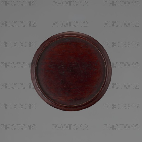 Vase on saucer. Cylindrical porcelain vase tapering toward the bottom with a flat rim. Light cream glaze with fine cracklework decoration. Wooden round flat saucer on three short legs. 19th century, Japan; Edo 1603–1868; Japanese ceramics. Height 21.1 cm; diameter 8.4 cm; weight 0.6 kg., A round wooden object with a dark reddish-brown color sits centered against a smooth gray background; the wood surface shows a few lighter and darker areas and several fine scratches, and the outer rim is slightly raised and darker than the central area.