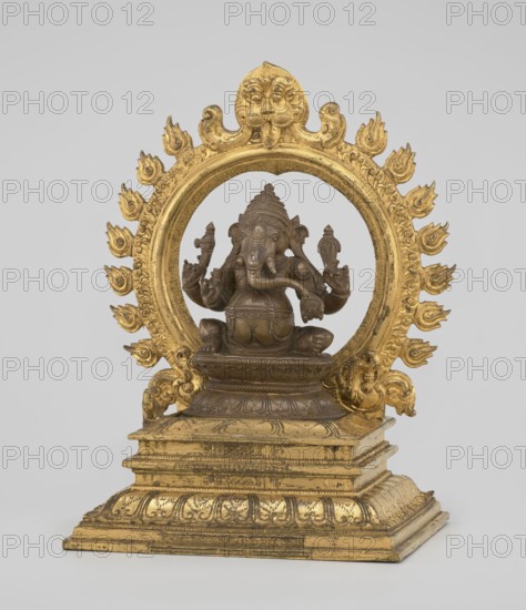 Ganesha, 19th century, India. Sculpture height 26.0 cm, width 17.5 cm. The deity sits on a double lotus throne placed on a rectangular flat base; the whole is mounted on a large, strongly profiled pedestal. At the back of the figure is a circle of fire. The figurine consists of three parts: a statuette inserted onto a pedestal and a fire ring inserted into the pedestal holes using two pins. Iconography: Indian/Hindu art — elephants, lotus throne, halos, birds, rats, fire, crowns/tiaras, tusks, mounts, armor; associated with Shaivism., A sculpted figure seated on a tiered rectangular base, framed by a decorative circular halo with flame-like motifs around its rim; the sculpture combines a darker bronze-like tone for the central figure with a brighter gold tone for the halo and base, showing detailed carved ornamentation and textured surfaces, with the figure posed with multiple raised arms and a trunk-like facial feature, wearing a tiered headdress and ornate adornments, and the halo to...