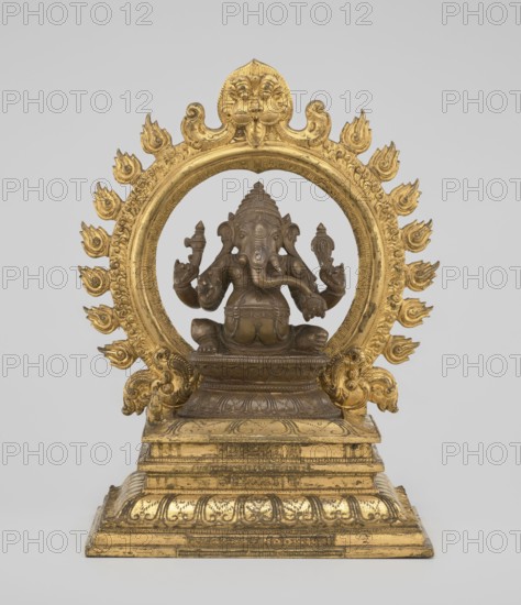 Ganesha, Indian, 19th century. Sculpture, height 26.0 cm, width 17.5 cm. The deity sits on a double lotus throne placed on a rectangular flat base; the whole is mounted on a large, strongly profiled pedestal. At the back of the figure is a circle of fire. The figurine consists of three parts: a statuette inserted onto a pedestal and a fire ring inserted into the pedestal holes using two pins. Iconography/subjects: Indian art, Hindu art, Hinduism, Shaivism; elephants, tusks, mounts, crowns/tiaras, halos, lotus throne, ropes, birds, rats, fire, armor, gluttony., A sculptural figure sits within an ornate circular frame mounted on a stepped rectangular base; the figure appears carved with multiple arms and an elephant-like head and rests on a decorated pedestal, all rendered in a dark brown tone, while the circular frame, ornamental flame-like motifs around it, and the stepped base are gilded in a bright gold color.
