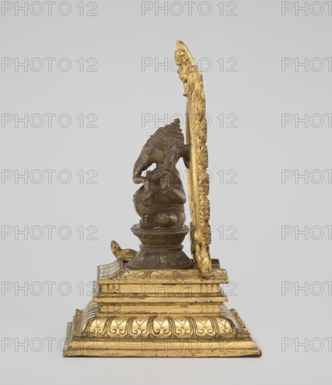 Ganesha, unknown sculptor. 19th century, India. Sculpture height 26.0 cm, width 17.5 cm. The deity sits on a double lotus throne placed on a rectangular flat base; the whole is mounted on a large, strongly profiled pedestal. At the back of the figure is a circle of fire. The figurine consists of three parts: a statuette inserted onto a pedestal and a fire ring inserted into the pedestal holes using two pins. Iconography/features: Indian/Hindu art; elephants, ropes, halos, lotus throne, birds, gluttony, rats, fire, Shaivism, crowns/tiaras, tusks, mounts, armor., A small sculpture of an elephant-headed figure seated on a rounded pedestal attached to a tiered rectangular base, with an ornate upright backplate behind it; the figure and base show a metallic brown tone while the base, backplate, and decorative details are gilded gold, and the background is light gray.