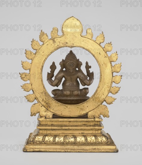 Ganesha, 19th century, India; sculpture; height 26.0 cm, width 17.5 cm. The deity sits on a double lotus throne placed on a rectangular flat base; the whole is mounted on a large, strongly profiled pedestal. At the back of the figure is a circle of fire. The figurine consists of three parts: a statuette inserted onto a pedestal and a fire ring inserted into the pedestal holes using two pins. Iconography: Indian/Hindu art — elephants, lotus throne, halos, birds, rats; attributes include crowns/tiaras, tusks, mounts, armor; associated with Hinduism and Shaivism., A gilded circular frame with a scalloped flame-like border surrounds a seated bronze figure viewed from the back; the frame and stepped base are gold with worn areas revealing darker tones, and the figure is a brownish bronze with detailed headgear, arm ornaments, and a seated posture on a small pedestal; the background is a pale gray.
