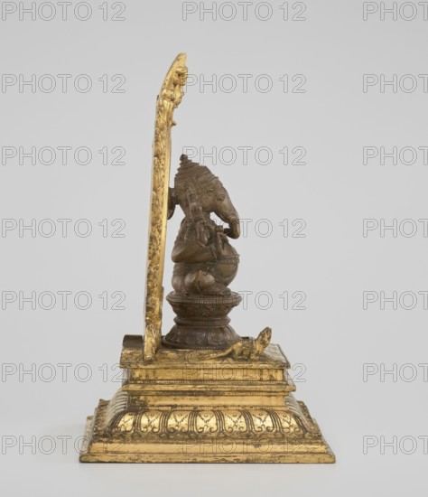 Ganesha, 19th century, India. Sculpture, height 26.0 cm, width 17.5 cm. The deity sits on a double lotus throne placed on a rectangular flat base; the whole is mounted on a large, strongly profiled pedestal. At the back of the figure is a circle of fire. The figurine consists of three parts: a statuette inserted onto a pedestal and a fire ring inserted into the pedestal holes using two pins. Iconography/details: Indian/Hindu art; Shaivism; elephants; tusks; crowns/tiaras; lotus throne; halos; ropes; mounts; armor; birds; rats; fire; gluttony; religion., A small sculptural object sits on a stepped rectangular base finished in gold tones with decorative relief patterns, supporting a vertical gold-toned backplate with carved ornamentation and a bronze-toned seated elephant-headed figure on a rounded pedestal; the figure faces to the right, has detailed headdress and body decoration, and rests on the pedestal which in turn sits on the gilt base, all set against a light gray background with...
