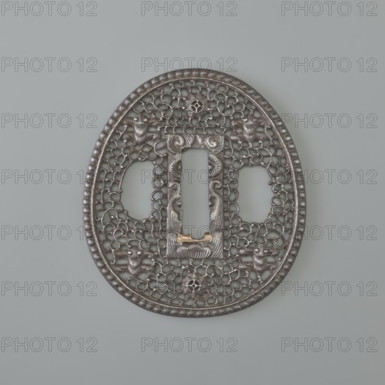 Crossguard tsuba, sword crossguard, namban style, openwork, flowers, openwork plant tendril karakusa with two sacred jewels tama woven into it and four mantis heads seen from the front. Egg-shaped tsuba with a separated edge and decorated with pearling; rectangular seppa dai decorated in low relief with a stylised volute ornament. Not signed. 18th century Edo period, 1603–1868, Japan. Tsuba; height 6.5 cm, width 5.8 cm, depth 0.35 cm., An oval metallic object with an openwork lattice pattern and a raised beaded rim, shown against a light gray background; the metal appears dark gray with areas of lighter gray and small touches of gold; the lattice contains a central rectangular opening flanked by two rounded openings and decorated with stylized animal or bird figures and floral-like motifs integrated into the filigree.