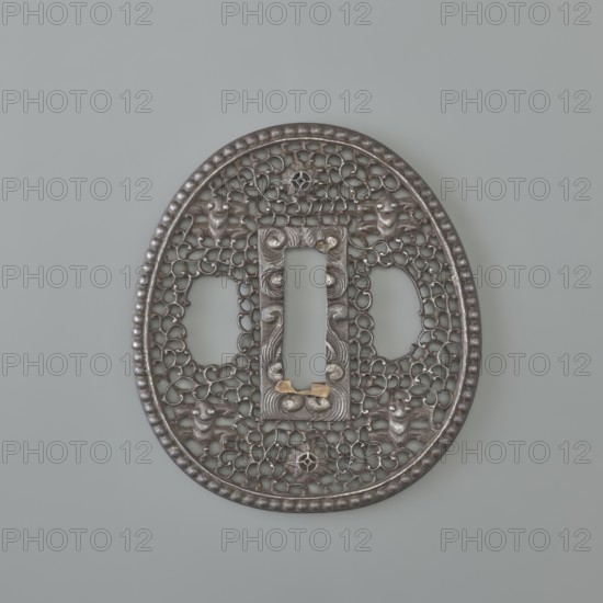 Crossguard tsuba, sword crossguard in namban style; openwork, egg-shaped tsuba with a separated edge and pearling. Rectangular seppa-dai, decorated in low relief with a stylised volute ornament. Surface decorated with an openwork plant tendril karakusa incorporating two sacred jewels tama and four mantis heads seen from the front. Not signed. 18th century Edo period, 1603–1868, Japan. Materials: metal tsuba; dimensions: height 6.5 cm, width 5.8 cm, depth 0.35 cm., A round, ornate metal object with an openwork lattice pattern sits against a smooth light gray background, showing a metallic surface in shades of dark gray and silver with small areas of muted gold. The object has a beaded outer rim and an inner rectangular opening framed by raised, carved details. A few symmetrical decorative motifs and flowing curves form a repeating network across the surface, and there are several rounded protrusions and textured elements integrated into the design. The overall color palette includes dar...