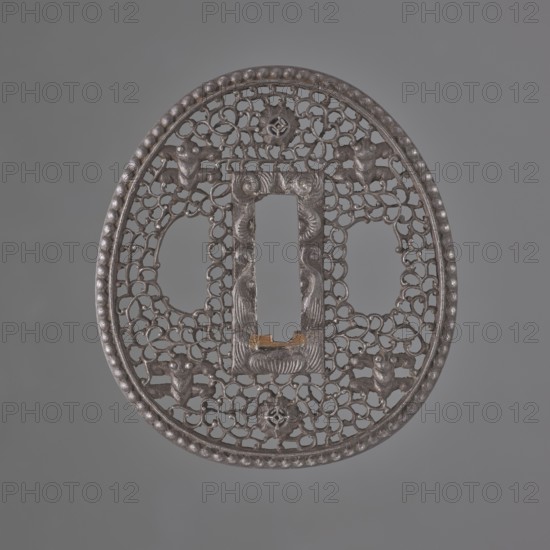 Crossguard tsuba, sword crossguard in namban style, openwork egg-shaped tsuba with a separated edge and pearling; rectangular seppa-dai decorated in low relief with a stylised volute ornament. Surface decorated with an openwork plant tendril karakusa with two sacred jewels tama woven into it and four mantis heads seen from the front. Not signed. 18th century Edo period, 1603–1868, Japan. Tsuba; height 6.5 cm, width 5.8 cm, depth 0.35 cm., A round, ornately pierced metal object with a raised beaded rim and an openwork lattice of interlocking curved shapes filling the interior; the central area features a vertically oriented rectangular opening framed by a carved border with symmetrical curved motifs and a small raised notch at its lower edge, and there are a few stylized animal-like figures integrated into the lattice around the central opening; the surface shows a metallic dark gray color with areas of slightly lighter gray highlights and a small patch of brownish-gold near the lower e...