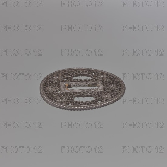 Crossguard tsuba, sword crossguard, namban style, openwork, flowers, tsuba, Japanese art, Japanese culture, openwork plant tendril karakusa with two sacred jewels tama woven into it and four mantis heads seen from the front. Tsuba of an egg shape, with a separated edge and decorated with pearling; rectangular seppa-dai decorated in low relief with a stylised volute ornament. Not signed. 18th century Edo period, 1603–1868, Japan. Materials: metal. Dimensions: height 6.5 cm, width 5.8 cm, depth 0.35 cm., A decorative round metal object sits against a smooth light gray background; the object is dark brown to black with some metallic sheen and small areas of lighter brown patina, and it has an ornate openwork pattern with floral and looping motifs and a beaded outer rim.