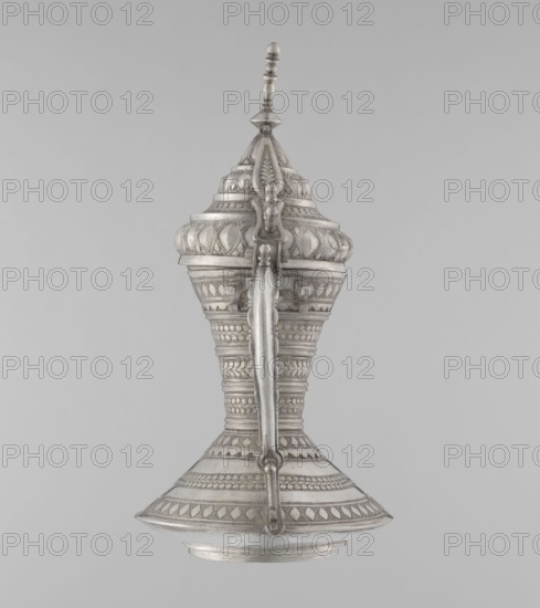 Teapot, Persian, Qajar dynasty 1785–1925. Silver teapot vessel with geometric motifs, used as a tea/coffee kettle. 19th–20th century, Iran. Weight: 580 g; height: 24 cm; width: 21.5 cm; diameter: 12.5 cm., The image shows a single ornate object centered against a plain light gray background; the object appears to be a tall, symmetrical vessel with a pointed finial at the top, a rounded bulbous midsection, a narrowing waist, and a flared base. The surface is covered in repeating decorative patterns and bands of carved or embossed motifs, including leaf-like shapes and geometric borders, and there is a prominent vertical handle running from near the top to the base. The visible colors are shades of silver and gray with subtle tonal variations where shadows and highlights accentuate the raised decorative details.