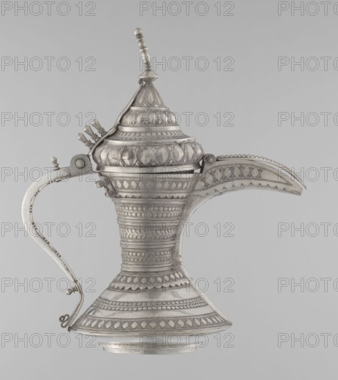 Teapot, Persian Iran, Qajar dynasty 1785–1925. 19th–20th century. Silver vessel for tea/coffee, geometric motifs. Height 24 cm; width 21.5 cm; diameter 12.5 cm; weight 580 g., A metal ewer with ornate raised patterns and a pointed lid topped by a finial, a curved spout with decorative edging, and an arched handle attached to the body; the surface shows embossed geometric and leaf-like motifs in shades of silver and gray against a light gray background.