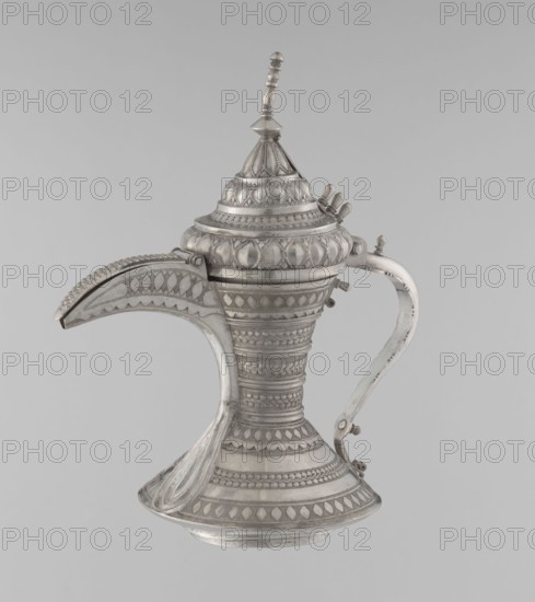 Teapot, Persian, Qajar dynasty 1785–1925, 19th–20th century. Iran. Brass vessel with geometric motifs; teapot/tea kettle for beverages tea/coffee. Height 24 cm; width 21.5 cm; diameter 12.5 cm; weight 580 g., A silver-colored ornate metal ewer with a pointed domed lid topped by a finial, featuring a curved spout and a separate curved handle; the body and lid are covered in repeating geometric and leaf-like raised patterns and bands, with decorative ridges and bead-like details, and the surface shows areas of smooth polish and slightly darker patina.