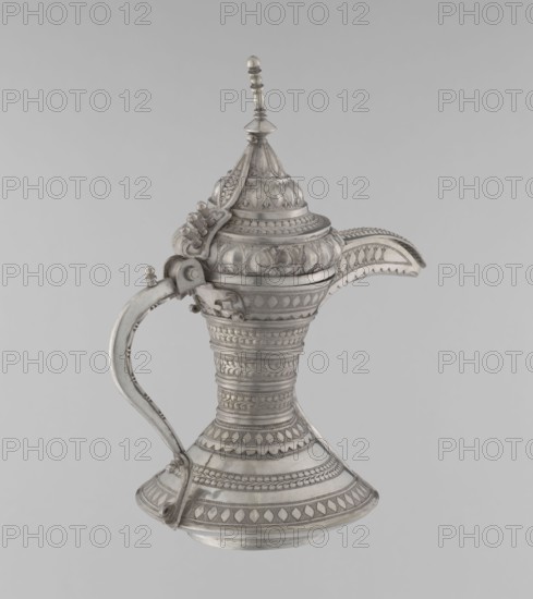 Teapot, Persian, Qajar dynasty 1785–1925; teapot/kettle for beverages tea/coffee; geometric motifs; 19th–20th century; Iran; vessel; weight 580 g; height 24 cm; width 21.5 cm; diameter 12.5 cm., A decorative metal vessel with an ornate lid and a curved spout is shown against a plain light gray background; the vessel features a tall, flared base that tapers to a narrow waist and widens again toward the top, a curved handle attached from near the base to the upper body, and a pointed, layered lid topped by a small finial; the surface is covered in repeating embossed patterns and textures including bands of geometric motifs and raised teardrop shapes; the visible colors are silver and shades of gray.