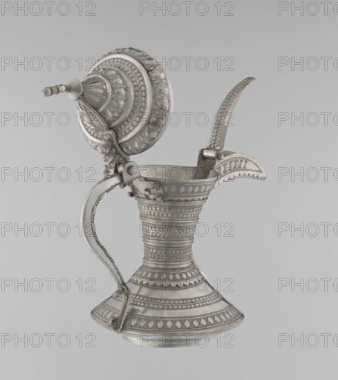 Teapot, Persian, Qajar dynasty 1785–1925. 19th–20th century Iran. Teapot/kettle for beverages tea/coffee, vessel with geometric motifs. Material not specified. Height 24 cm; width 21.5 cm; diameter 12.5 cm; weight 580 g., A metal pitcher with a hinged domed lid and a curved handle sits against a neutral background; the pitcher is primarily silver in color with darker gray patina in recesses, and features intricate raised decorative bands and repeating geometric and leaf-like patterns covering the body, neck, spout, base, lid, and handle. The lid has concentric circular ornamentation and a tapered finial extending outward, while the spout has a scalloped edge and textured top. The handle is attached with visible hinge fittings and shows similar patterned detailing. The background is a uniform light gray.