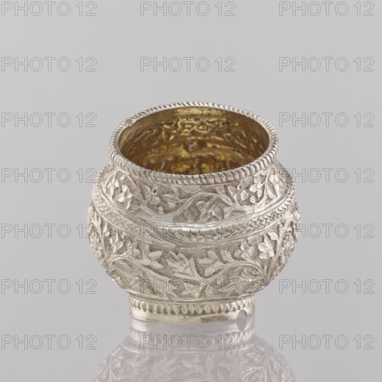 Bowl; maker, 19th century, India; Indian art; vessel; applied art; plant ornament with flowers and leaves; cup/container; height 4.5 cm; diameter 4.0 cm; weight 50.0 g., A small ornate rounded metal vessel sits on a reflective surface against a light gray background; the exterior of the vessel shows intricate raised floral and vine patterns in a silvery color with a slightly darker patina in recesses, and the interior appears gold-toned; the rim has a twisted rope-like border and the base shows a decorative band and a small notch; visible colors include silver, gold, gray, and hints of darker shadows.