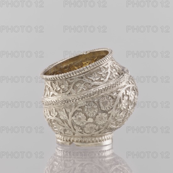 Bowl, 19th century India, applied art vessel; materials/weights: 50.0 g; dimensions: height 4.5 cm, diameter 4.0 cm. Decoration: plant ornament—flowers and leaves; function: cup/container., A small ornate metal vessel with a rounded, slightly angled opening; the surface is covered in detailed raised floral and vine patterns and textured bands, and the rim has a ridged edge. The exterior appears silver in color with reflective highlights, while the interior shows a warmer, golden color. The object sits on a reflective surface and is set against a plain light gray background.