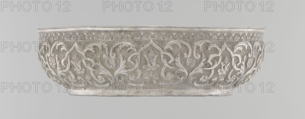 Bowl, purchase provenance noted, Islamic art, Turkish, Ottoman dynasty 1299–1922, geometric motifs and plant ornament, tughra of Abdulhamid II Ottoman sultan; 1842–1918, dated 1876–1909, Turkey, silver hammam vessel, height 5.6 cm, diameter 19.0 cm, A shallow, oval metal bowl with ornate raised floral and foliate relief work across its curved sides, featuring flowing scrolls and stylized leaf and flower motifs around the exterior; the rim has a narrow decorative band with repeating small patterns, and the base shows a simple flattened foot. The visible colors are silver and gray tones with slight variations in sheen and patina.