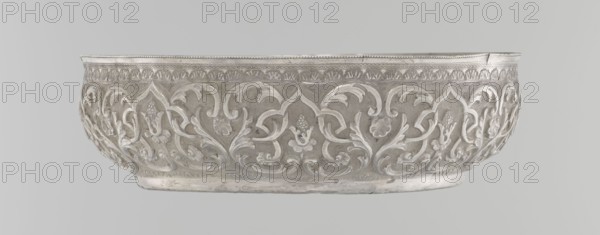 Bowl, Islamic art, Turkish, Ottoman dynasty 1299–1922, geometric motifs and plant ornament, tugra of Abdulhamid II 1842–1918, silver hammam bowl, dated 1876–1909, Turkey; vessel, height 5.6 cm, diameter 19.0 cm., A wide shallow bowl with ornate raised floral and foliate decorations covering its exterior surface, featuring flowing vine-like scrolls and leaf motifs in a repeating pattern; the rim has a narrow decorative band with a beaded edge and a carved frieze beneath it, and the base shows a plain slightly rounded foot. The visible colors are silver, light gray, and darker gray tones.