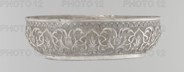 Bowl, Islamic art, Turkish, Ottoman dynasty 1299–1922, geometric motifs, plant ornament, tugra of Abdulhamid II Ottoman sultan; 1842–1918, 1876–1909, Turkey; vessel; silver hammam bowl; height 5.6 cm, diameter 19.0 cm., A shallow oval bowl with ornate raised floral and foliate relief decoration around its exterior, showing a repeating pattern of stylized leaves and flower motifs; the rim has a band of small decorative beading and a narrow patterned border beneath it; the surface appears metallic with areas of higher polish and some darker patina, showing shades of silver and gray; the background behind the bowl is a uniform pale gray.