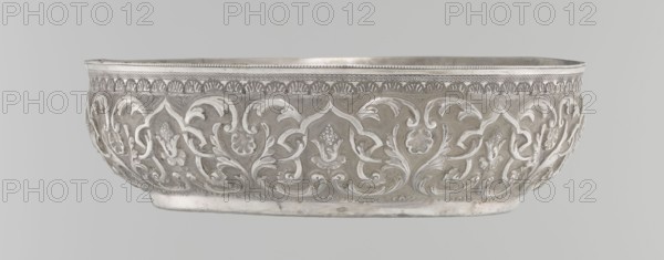 Bowl, purchased provenance noted, Islamic art, Turkish, Ottoman dynasty 1299–1922, geometric motifs and plant ornament, tugra of Abdulhamid II Ottoman sultan; 1842–1918, analogy noted, dated 1876–1909, Turkey, vessel, height 5.6 cm, diameter 19.0 cm, A shallow, oval metal bowl with an ornate raised floral and foliate pattern wrapping around its exterior, showing curved leaf motifs and stylized blossoms in relief; the rim features a patterned border with a small beaded edge and repeated decorative elements beneath it; the bowl rests on a slightly raised base and has a smooth interior visible at the top; the visible colors are shades of silver and gray with subtle variations in tone.