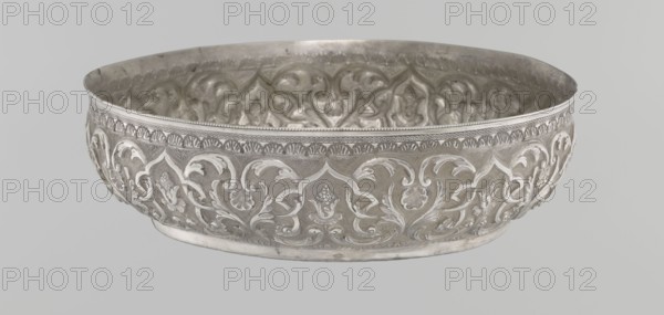 Bowl, Islamic art, Turkish, Ottoman dynasty 1299–1922, geometric motifs, plant ornament, tugra of Abdulhamid II Ottoman sultan; 1842–1918, 1876–1909, Turkey; vessel; silver hammam bowl; height 5.6 cm, diameter 19.0 cm, A shallow decorative metal bowl with an embossed floral and scroll pattern around its exterior and interior, showing raised curving vine-like motifs and petal shapes; the rim has a narrow band of repeating small ornamental shapes. The surface appears metallic with shades of silver and gray, and the background is a light gray.